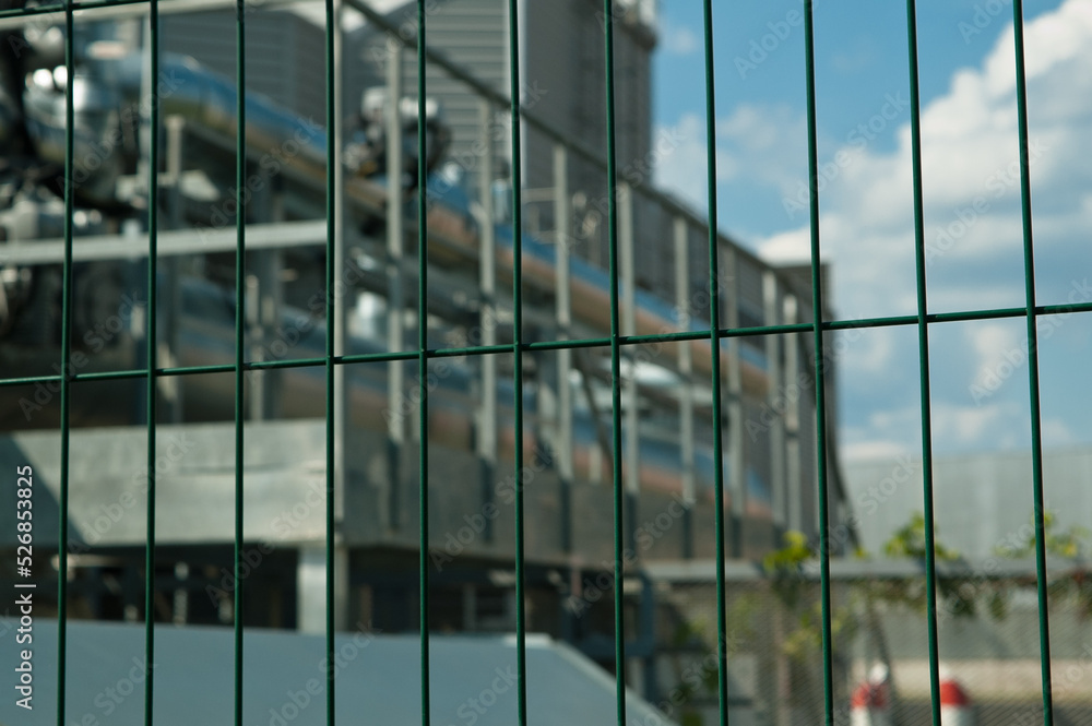 Obraz premium metal 3D mesh, in the photo a mesh fence in the background is a power plant building and a blue sky