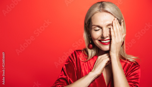 Beautiful middle aged woman with blond hair, stylish makeup, lauging and smiling, touching her flawless skin, standing over red background