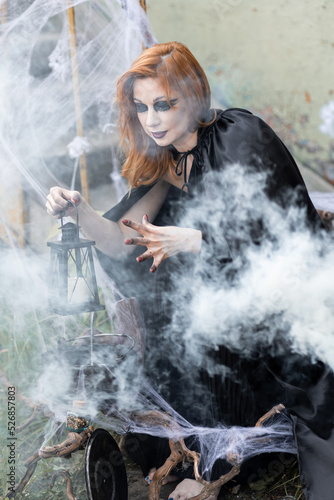 Attractive beautiful woman in a Halloween costume black cloak and scary make-up. Young girl in a look of a witch dressed up for a party making fairy potion holding black lantern