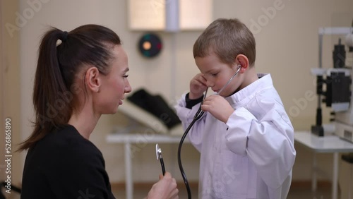 Positive woman passing stethoscope to boy listening auscultation standing in hospital indoors. Side view of Caucasian mother and son choosing future profession talking smiling photo