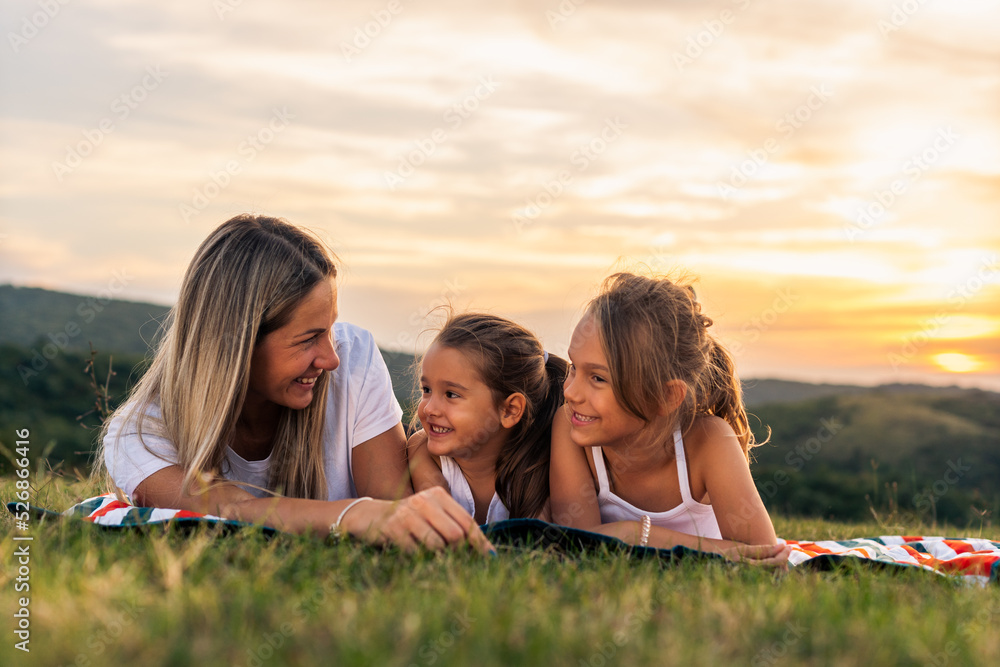 Happy mother and daughters smiling and laughing lying on blanket at the lawn in summer park. Family spend their free time together on vacation. Happy mother and daughters smiling and laughing lying on blanket at the lawn in summer park. Family spend their free time together on vacation.