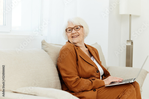 granny sat down on the sofa in a stylish brown suit and looks sweetly into the camera smiling benevolently, holding a laptop on her lap typing text © Tatiana