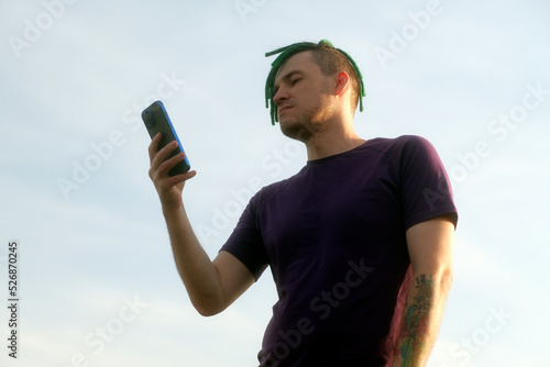 Portrait of young man with dreadlocks browsing mobile phone on background of cloudy sky.