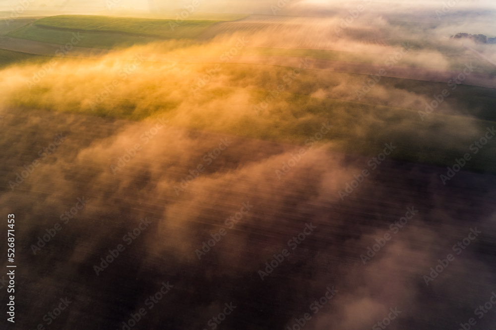 Fototapeta premium Thick fog covering colorful farm fields with orange glaze from rising sun. Morning landscape of Roztocze Poland. Birdseye view. Horizontal shot. High quality photo