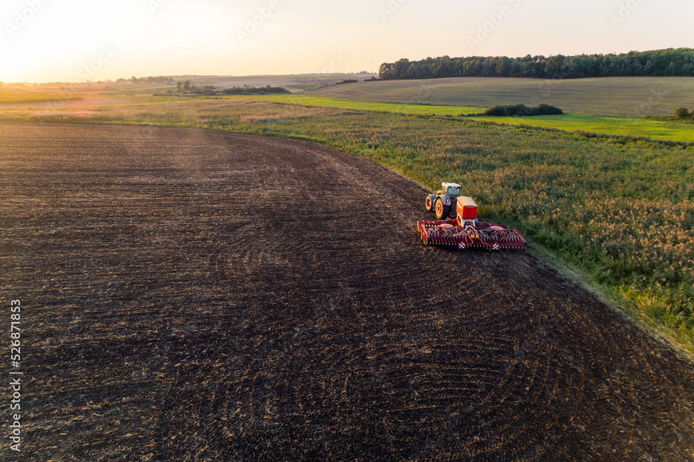 Obraz premium Tractor with sowing machine driving through brown field with greenery on the side and in the fading background. Morning landscape. Horizontal shot. High quality photo
