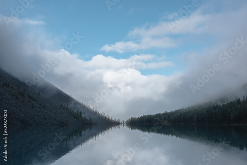 Tranquil meditative misty scenery of glacial lake with reflection of pointy fir tops and clouds at early morning. Graphic EQ of spruce silhouettes on calm alpine lake horizon. Mountain lake in fog.