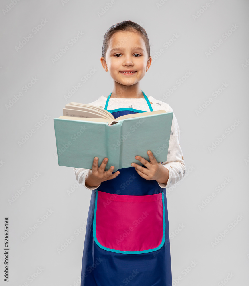 cooking, culinary and profession concept - happy smiling little girl in apron reading cook book over grey background cooking, culinary and profession concept - happy smiling little girl in apron reading cook book over grey background