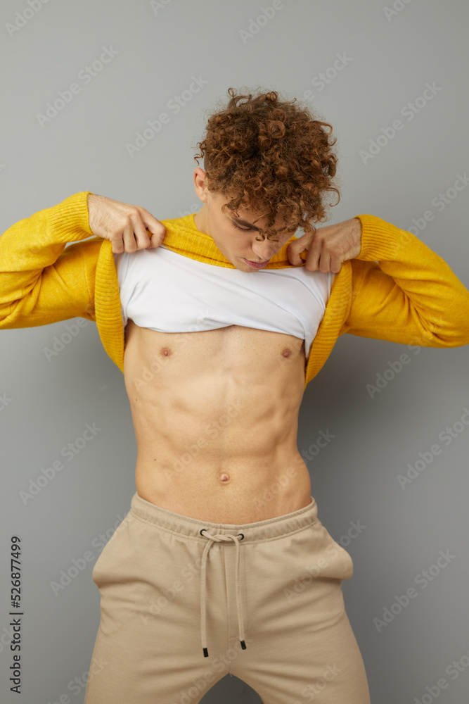 vertical photo on an isolated gray background of a handsome young man examining his perfect abs by lifting an orange sweater