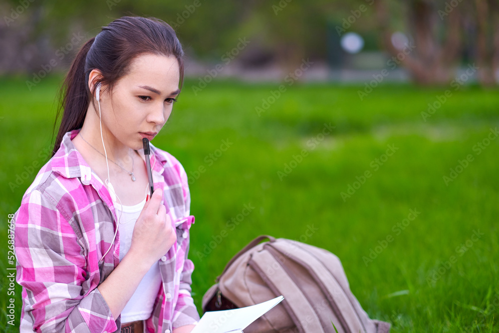 Young Asian woman Student with a book, notebook and pen on grass in summer park. Preparation from session and exams. outdoor portrait Young Asian woman Student with a book, notebook and pen on grass in summer park. Preparation from session and exams. outdoor portrait