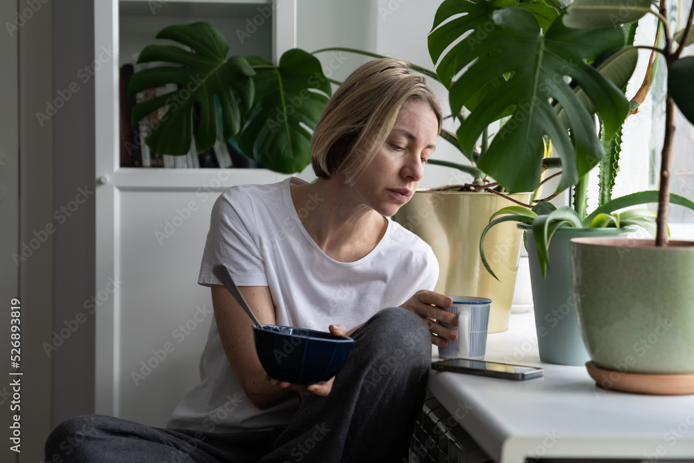 Upset single middle-aged female thinks about uneasy life taking mug with hot drink sitting near window. Blonde woman holds breakfast bowl searching job via smartphone put on windowsill closeup Upset single middle-aged female thinks about uneasy life taking mug with hot drink sitting near window. Blonde woman holds breakfast bowl searching job via smartphone put on windowsill closeup