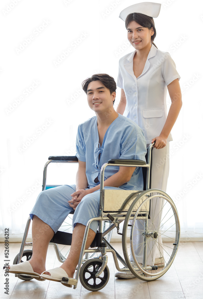 Asian young professional cheerful female assistant nurse in white uniform help pushing happy healthy male patient in blue hospital clothes sitting on wheelchair smiling after recovery in hallway Asian young professional cheerful female assistant nurse in white uniform help pushing happy healthy male patient in blue hospital clothes sitting on wheelchair smiling after recovery in hallway