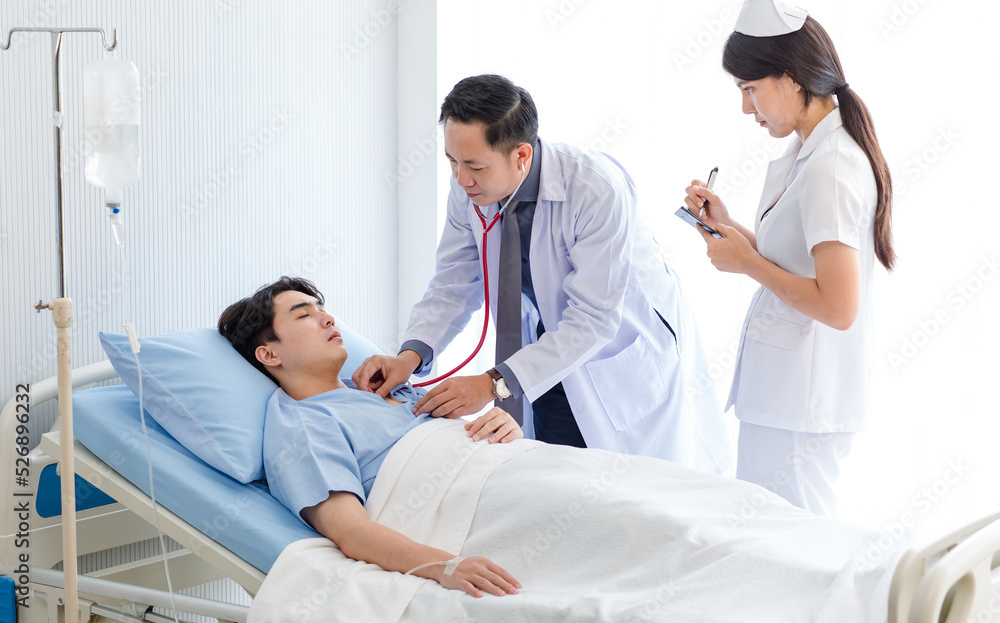 Asian professional male doctor in white lab coat using stethoscope listening monitoring heartbeat pulse on patient chest while female nurse in uniform writing symptom data on clipboard in ward room Asian professional male doctor in white lab coat using stethoscope listening monitoring heartbeat pulse on patient chest while female nurse in uniform writing symptom data on clipboard in ward room