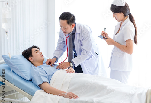 Asian professional male doctor in white lab coat using stethoscope listening monitoring heartbeat pulse on patient chest while female nurse in uniform writing symptom data on clipboard in ward room