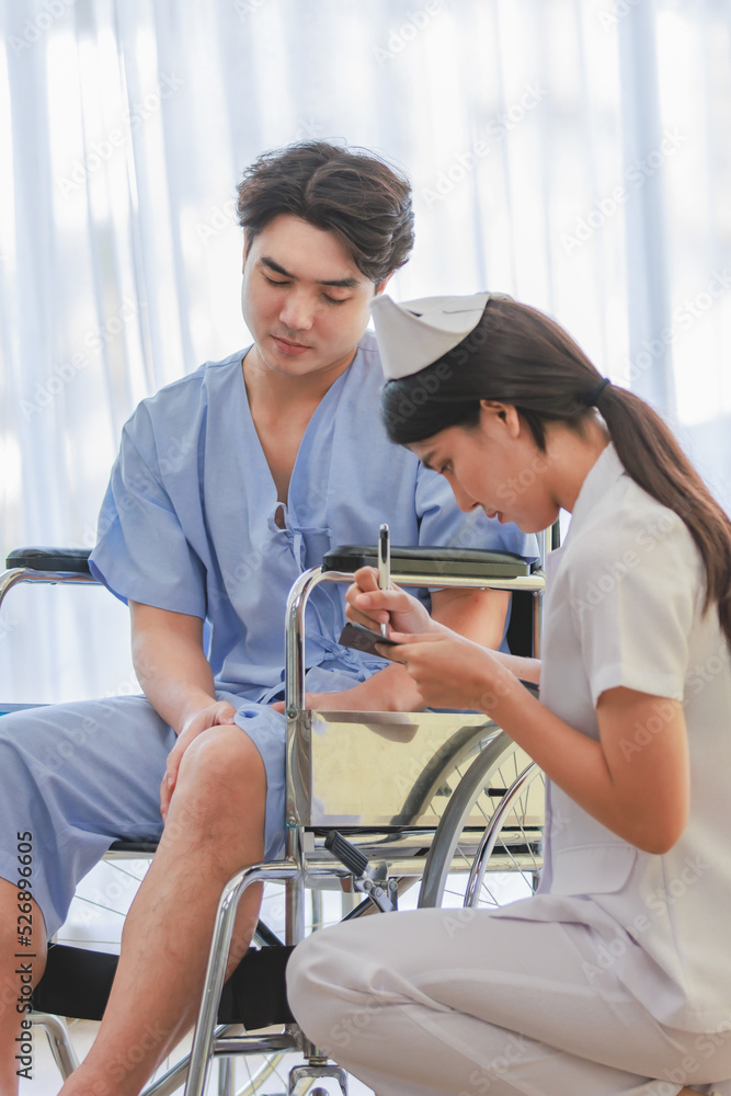 Obraz premium Asian professional young female nurse in white uniform holding clipboard writing note on paper while visiting monitoring asking male patient in blue hospital clothes sitting on wheelchair in ward