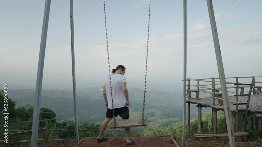 Man walks and sits down on swing by green hilly landscape in Thailand Man walks and sits down on swing by green hilly landscape in Thailand