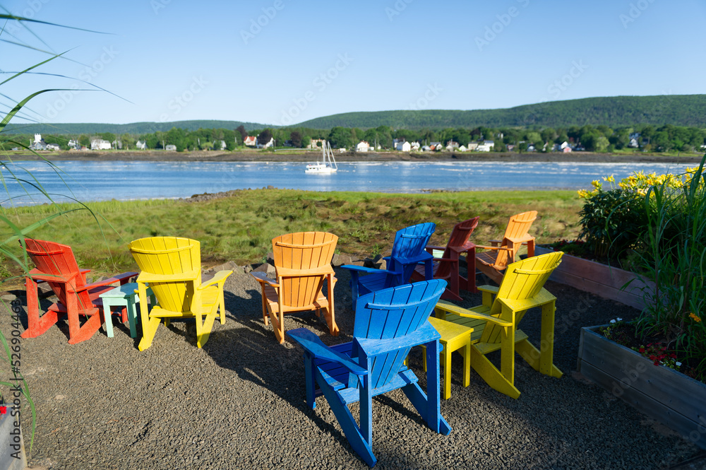 Multicolor Adirondack chairs facing the calm water of the Bay of Fundy in Nova Scotia, Canada on a beautiful summer day