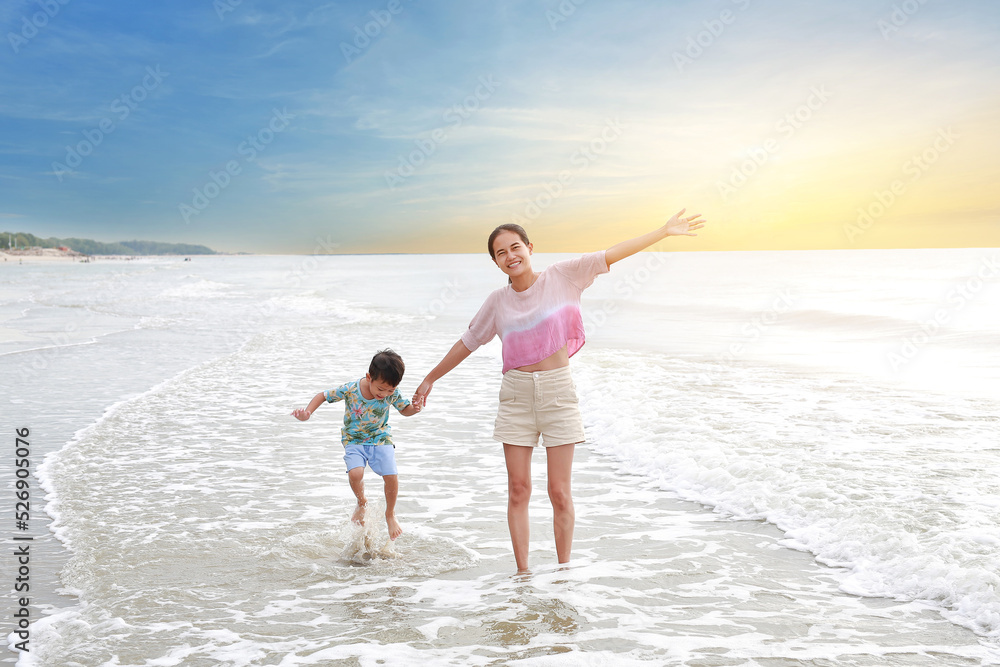 Cheerful Asian mother and little boy enjoy playing on tropical sand beach at sunrise. Happy family mom and son having fun in summer holiday.