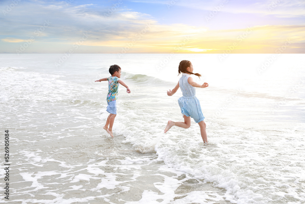 Cheerful Asian young sister and little brother having fun together on tropical sand beach at sunrise. Happy family boy and girl enjoy in summer holiday.