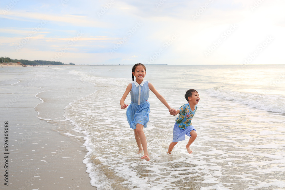Cheerful Asian child girl and little boy having fun run together and hand in hands on tropical sand beach at sunset. Happy family enjoy in summer holiday.