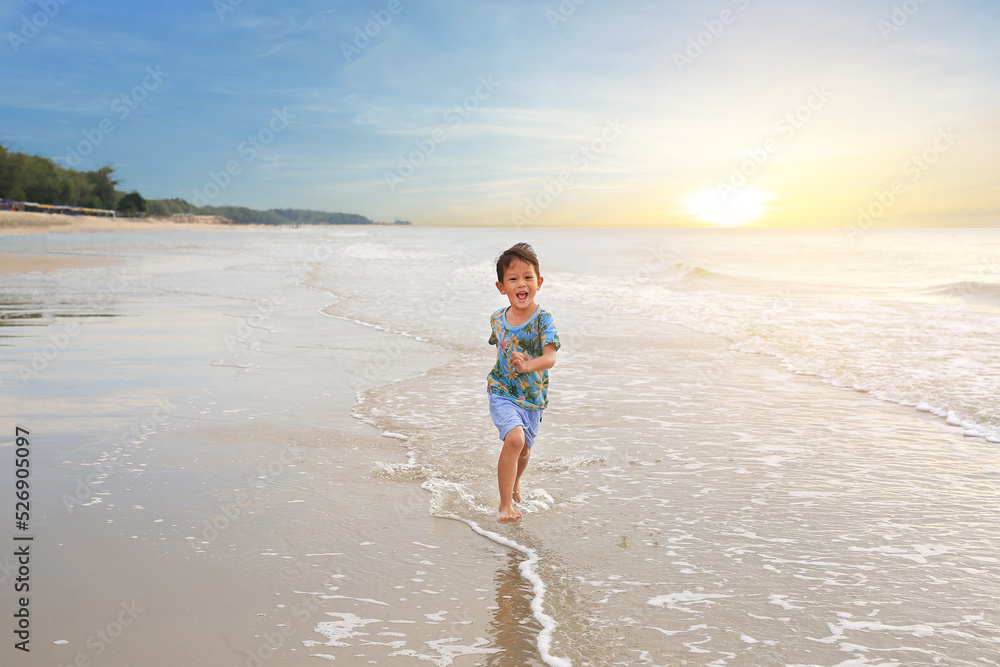 Happy Asian little boy having fun running on tropical sand beach at sunrise.