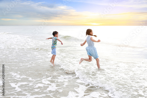 Cheerful Asian young sister and little brother having fun together on tropical sand beach at sunrise. Happy family boy and girl enjoy in summer holiday.