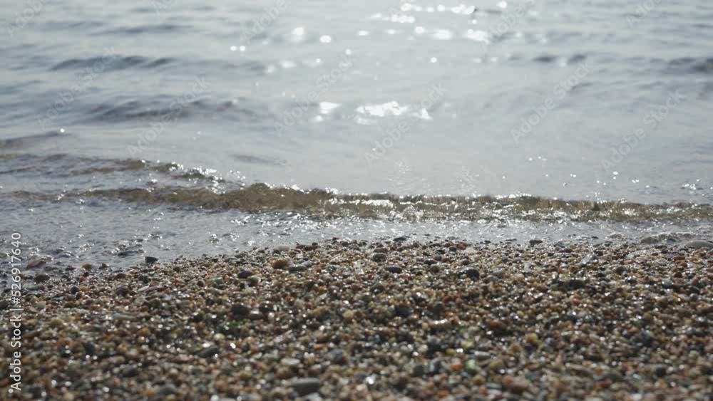 Close-up of pebble beach with transparent waves with sun glare on the water. Close up of stony sea shore. Natual background of calm sea in the surf zone. Camera moves sideway to the left side Close-up of pebble beach with transparent waves with sun glare on the water. Close up of stony sea shore. Natual background of calm sea in the surf zone. Camera moves sideway to the left side