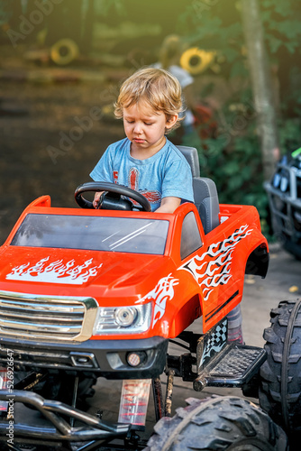 Frustrated toddler boy in blue t-shirt sits in red children car with big wheels in public park in summer evening
