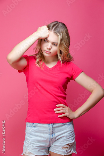 One Beautiful Sad Depressed Caucasian Girl in Pink T-Shirt Posing in Summer Shorts With Lifted Hand Thinking Against Coral Pink Background.