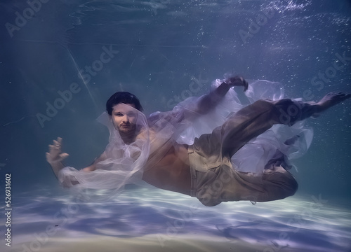young man in white ribbons and trousers underwater in the pool on a dark background