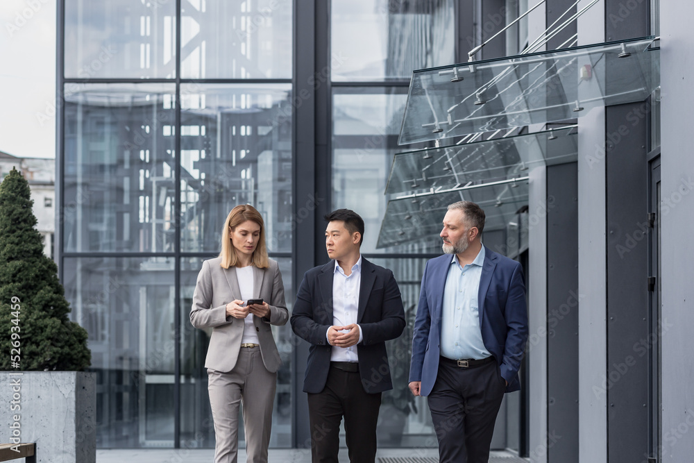 Diverse business group, three male and female workers walking and chatting discussing plans, outside office building Diverse business group, three male and female workers walking and chatting discussing plans, outside office building