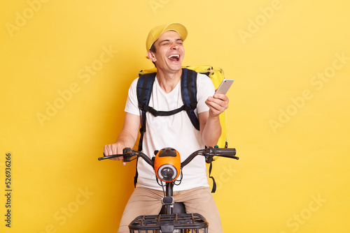 Indoor shot of happy young delivery guy holding cell phone, wearing cap and thermo bag, riding bicycle, laughing, expressing positive emotions, isolated on yellow background
