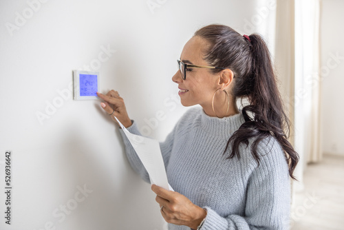 Smiling woman operates digital thermostat installed on the wall inside her home photo