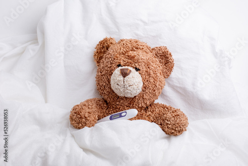 teddy bear is lying in bed under a blanket with a thermometer, white background, the concept of health and medicine
