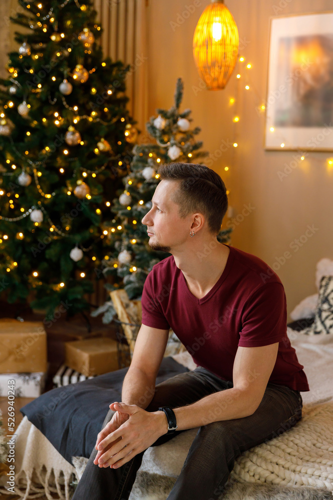 brunet man in T-shirt and jeans sits on bed in room decorated for Christmas brunet man in T-shirt and jeans sits on bed in room decorated for Christmas