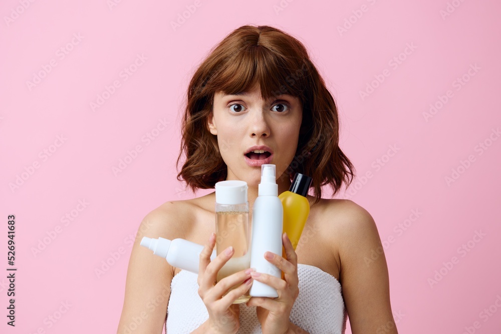 a young, pleasantly surprised woman stands on a pink background holding different jars of care cosmetics in her hands and happily looks into the camera with her mouth wide open. a young, pleasantly surprised woman stands on a pink background holding different jars of care cosmetics in her hands and happily looks into the camera with her mouth wide open.