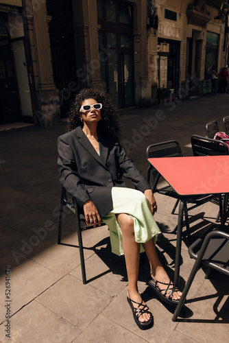 Calm young african woman sittin on chair in cafe spends free time outdoor in sunny spring day. Brunette wears dress, jacket and sunglasses. Concept relaxing time.