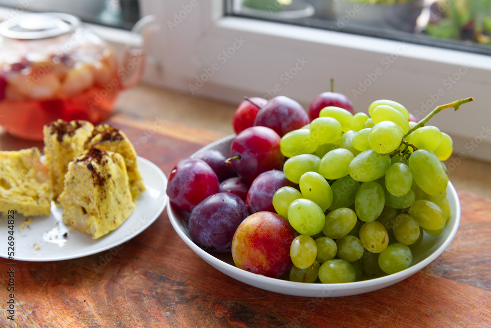 slices of homemade cake on a plate, green grape and plums, a glass teapot with stewed fruits on a kitchen windowsill, concept of fresh fruits and healthy food slices of homemade cake on a plate, green grape and plums, a glass teapot with stewed fruits on a kitchen windowsill, concept of fresh fruits and healthy food