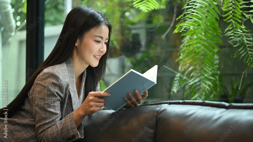 Fototapeta premium Elegant millennial business woman resting on couch in bright modern office and reading book