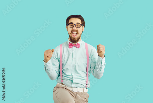 Cheerful happy male hipster nerd rejoices in his success standing on light blue background. Guy with mustache and beard wearing shirt, suspenders and glasses happily clenched his fists shouting YES.