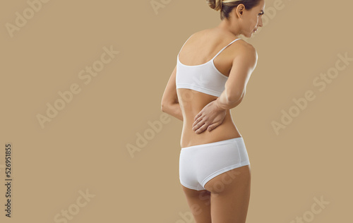 Backside view from behind woman in underwear isolated on beige color text background suffering from pain and holding hand on low back area. Body injury, back pain, renal cancer, kidney disease concept © Studio Romantic