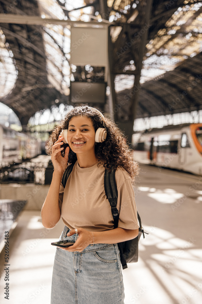 Smiling young african woman with headphones and smartphone listening music on railway station. Brunette wears casual clothes and backpack. Concept enjoying travel, music part of life.