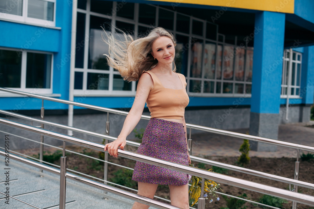A beautiful girl with blond long hair, dressed in a bright skirt and a sexy topic, holds a bouquet of wild flowers in her hands, against the backdrop of a modern building.