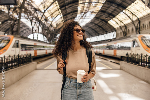 Happy young african female in sunglasses looking into distance smiling with teeth holds coffee. Brunette wears light brown t-shirt and jeans skirt. Concept positive moments, traveler.