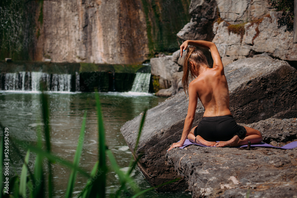 Fototapeta premium A beautiful tanned girl with a sports figure is doing yoga exercises against the backdrop of a picturesque waterfall in the summer. Sits in the lotus position. She is wearing black shorts and a top.