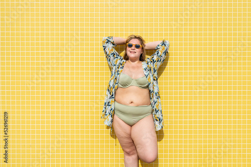 Smiling plus size woman with hands behind head in front of yellow wall photo