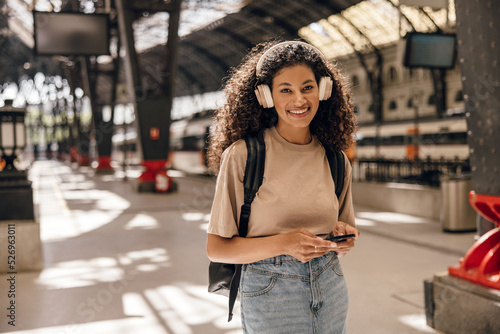 Canvas-taulu Smiling young african woman looking at camera with good mood uses her smartphone on railway station