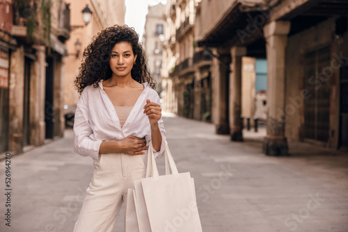 Stylish young african girl looking into camera spends free time on street in sunny weather. Brunette wears singlet, shirt and white jeans. Concept shopping, walking alone.