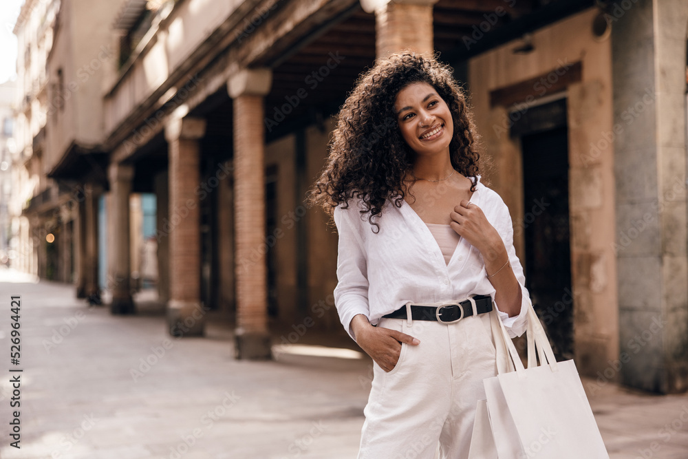 Pretty young african woman with curly hair looks at distance smiling teeth on old block. Model brunette hair wears singlet, shirt and white jeans. Concept fashionable vacation.