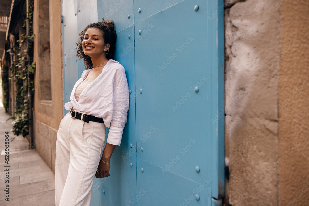 Positive young african woman smiling into camera, stands at europe street near blue gate. Brunette wears singlet, shirt and casual white jeans. Concept leisure vacation.