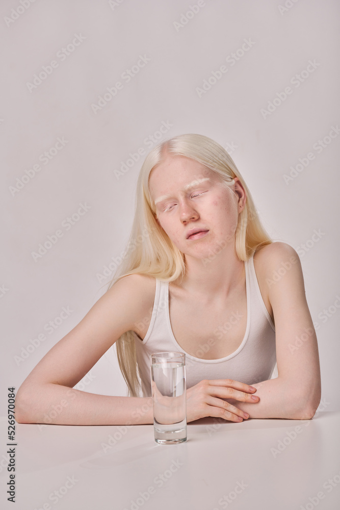 Young albino girl with white long hair sitting at table with glass of water on white background - obrazy, fototapety, plakaty 